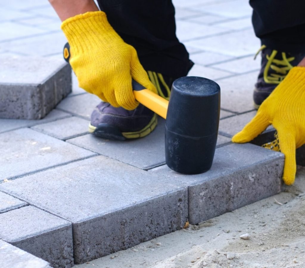 The master in yellow gloves lays paving stones in layers. Garden brick pathway paving by professional paver worker. Laying gray concrete paving slabs in house courtyard on sand foundation base.